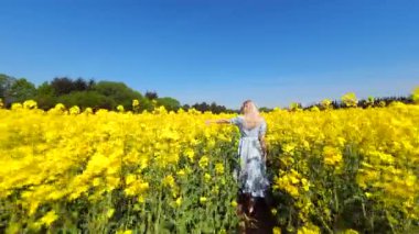 Güneşli bahar gününde sarı kolza tohumu tarlasında yürüyen bir kadının sinematik FPV görüntüsü. Doğa, özgürlük ve yavaş yaşam temaları için huzurlu kırsal yaşam tarzı sahneleri.