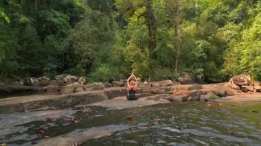 Huzurlu tropikal ormanda yoğun yeşilliklerle çevrili kayalar üzerinde meditasyon yapan bir kadın. Sağlık, farkındalık ve seyahat temaları için ideal.
