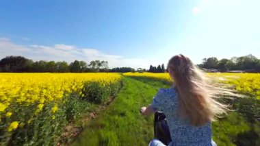 Güneşli bir günde sarı kolza tohumu tarlasında motosiklet süren bir kadının sinematik FPV görüntüsü. Özgürlük ve yaz temaları için enerjik kırsal yaşam tarzı manzarası.