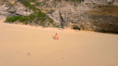 FPV 'nin uzaktan kumsalda tek başına meditasyon yapan kadın görüntüsü. Turkuaz deniz, dalgalar ve uçurumlar, Bali, Endonezya. Rahatlamanın, farkındalığın ve iç huzurun huzurlu sahnesi..
