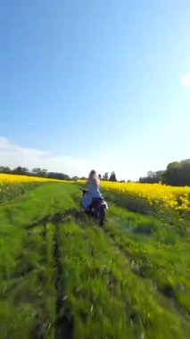 Güneşli bir günde sarı kolza tohumu tarlasında motosiklet süren bir kadının sinematik FPV görüntüsü. Özgürlük ve yaz temaları için enerjik kırsal yaşam tarzı manzarası.