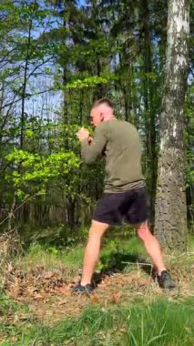 Atletik adam yeşil ormanda dövüş sanatlarını eğitiyor, diz vuruşları ve boks hareketleri yapıyor. Doğada güç ve disiplin.