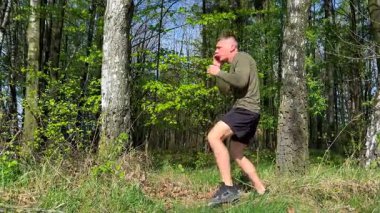 Atletik adam yeşil ormanda dövüş sanatlarını eğitiyor, diz vuruşları ve boks hareketleri yapıyor. Doğada güç ve disiplin.