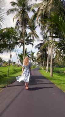 Endonezya, Bali 'de palmiye çizgili yolda çıplak ayaklı bir kadının uçuşan elbisesi. Pirinç tarlaları, tropikal titreşimler ve yavaş yaşam cazibesiyle huzurlu bir kır manzarası..