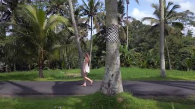 Endonezya, Bali 'de tropikal palmiye çizgili yolda yürüyen beyaz elbiseli bir kadın. Bereketli yeşillik, palmiye ağaçları ve sakin atmosferi olan huzurlu bir ada..