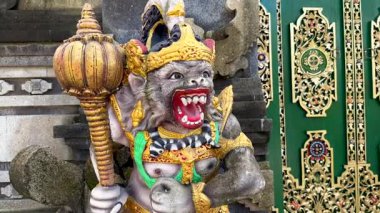 Endonezya, Bali 'deki tapınak girişini koruyan renkli Hanuman heykelinin canlı yakın çekimi. Altın tacı ve topuzu olan Hindu maymun tanrısı süslü yeşil tapınak kapılarının önünde duruyor..