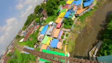 Dinamik FPV insansız hava aracı Malang, Endonezya 'daki renkli bir köy üzerinde uçuyor canlı gökkuşağı evleri, nehir kenarındaki evler ve yukarıdan gelen tropikal yaşam sergiliyor..
