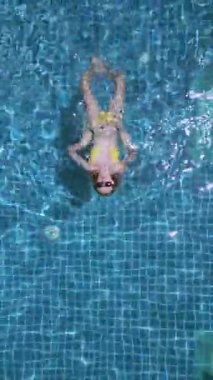 Açık mavi suyla tropikal havuzda yüzen sarı bikinili kadının yukarıdan aşağı görüntüsü. Rahatlama, yaz yaşam tarzı ve seyahat içeriği için mükemmel..