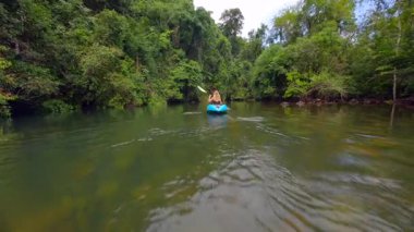 Tayland 'da yemyeşil bir ormanda tropikal nehirde kayak yapan bir kadının FPV görüntüsü. Huzurlu, sinematik doğa manzarası seyahat, eko-turizm ve açık hava yaşam tarzı projeleri için ideal.