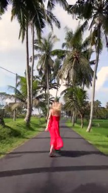 Kırmızı elbiseli neşeli kadın Endonezya, Bali 'de yeşil pirinç tarlalarıyla çevrili palmiyeli tropikal yolda neşe içinde atlıyor. Mutluluk, özgürlük ve doğada yavaş seyahat kavramı..