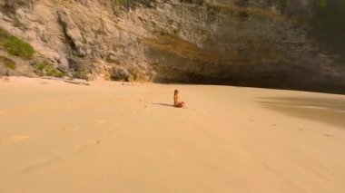 FPV 'nin uzaktan kumsalda tek başına meditasyon yapan kadın görüntüsü. Turkuaz deniz, dalgalar ve uçurumlar, Bali, Endonezya. Huzurlu bir rahatlama, farkındalık ve iyileşme sahnesi..