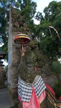 Bali, Endonezya 'da seremoni kapısı, taş heykeller, penjor direkleri ve kutsal kumaşla uçan FPV insansız hava aracı. Ruhaniyete ve kültüre sembolik yolculuk.