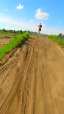 Motokros sürücüsünün kırsaldaki kumlu toprak pistte sıçrayışını ve sürüşünü takiben yavaş çekim FPV insansız hava aracı videosu