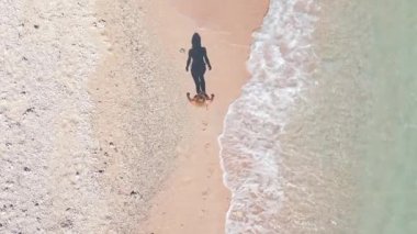 Kumlu tropik plajda kristal berrak deniz ve gölgesi kıyı şeridi boyunca yürüyen kadının yukarıdan aşağı görüntüsü. Yaz, tatil ve kıyı yaşam tarzının minimalist ve sanatsal sahnesi.