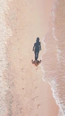 Kumlu tropik plajda kristal berrak deniz ve gölgesi kıyı şeridi boyunca yürüyen kadının yukarıdan aşağı görüntüsü. Yaz, tatil ve kıyı yaşam tarzının minimalist ve sanatsal sahnesi.