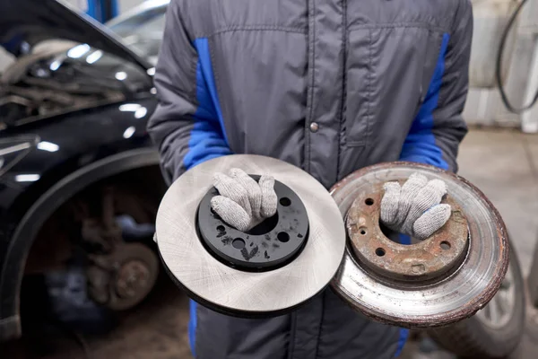 The mechanic holds old rusty brake disc and new disc. Change the old to new brake disc on car in a garage. Auto repair concept