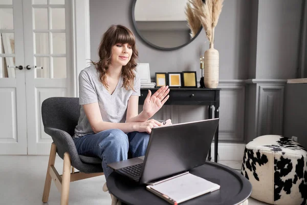 Woman waving hand has video call laptop computer online sitting in armchair at home. Hello there. Pleasant young female teacher or tutor sitting at home in cozy room before laptop. Hight