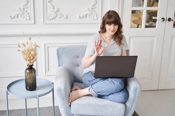 Woman waving hand has video call laptop computer online sitting in armchair at home. Hello there. Pleasant young female teacher or tutor sitting at home in cozy room before laptop. Hight