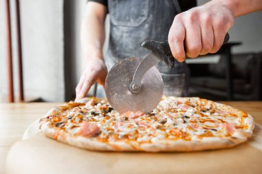 Closeup hand of chef baker in uniform blue apron cutting pizza at kitchen. High quality photo