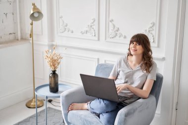 Evdeki oturma odasında kanepede otururken siyah dizüstü bilgisayar kullanan güzel, neşeli bir kız. Genç bir kadın iletişim ve iş için dizüstü bilgisayar kullanıyor. Evet.