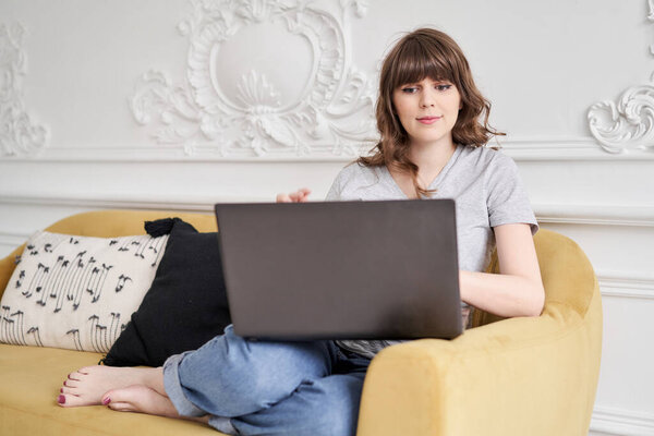 Beautiful cheerful girl using black laptop while sitting on sofa in living room at home. Young casual woman using laptop for communication and work. S
