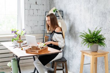 Genç bayan serbest çalışan dizüstü bilgisayarda çalışıyor, sosyal ağlardaki mesajlara cevap veriyor. Çiçek teslimatı konsepti. Çiçekçide profesyonel işçi.