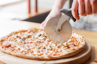 Closeup hand of chef baker in uniform blue apron cutting pizza at kitchen. High quality photo