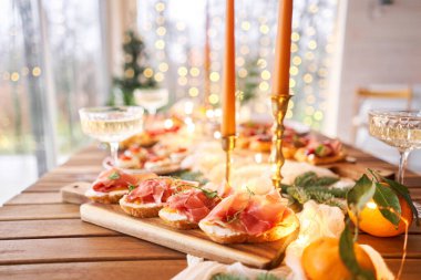 Noel yemeği ziyafeti. Küçük bir masa aperatifler, bruschetalar ve kanepelerle servis edilir. Şampanya bardakları, mumlar ve Noel ağacıyla süslü bir yemek masası. Arka planda bir çelenk..