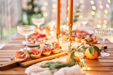 Noel yemeği ziyafeti. Küçük bir masa aperatifler, bruschetalar ve kanepelerle servis edilir. Şampanya bardakları, mumlar ve Noel ağacıyla süslü bir yemek masası. Arka planda bir çelenk..