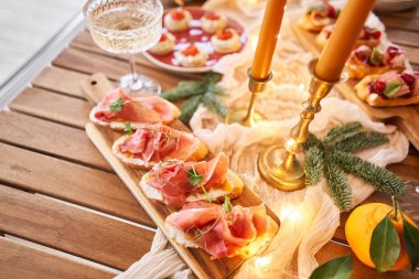 Noel yemeği ziyafeti. Küçük bir masa aperatifler, bruschetalar ve kanepelerle servis edilir. Şampanya bardakları, mumlar ve Noel ağacıyla süslü bir yemek masası. Arka planda bir çelenk..