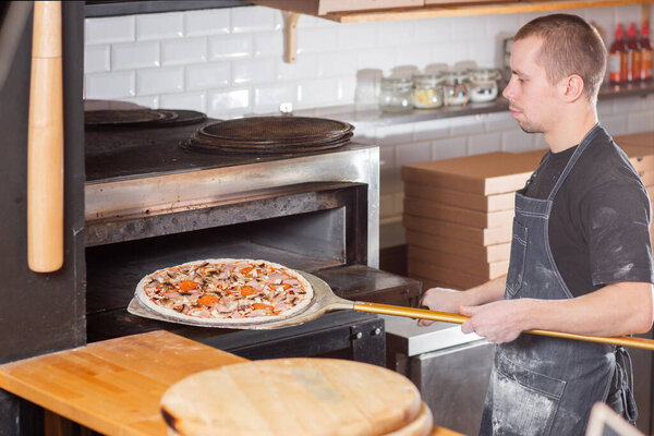 Closeup hand of chef baker in uniform blue apron cutting pizza at kitchen. High quality photo