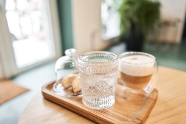 Bir fincan cantuccini ve ahşap tepside bir bardak su. Sütlü kahve, kurabiye ve cam sehpanın üzerinde su olan bir bardak. Yüksek kalite fotoğraf