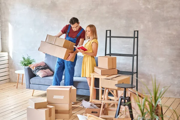 Delivery and moving day concept. The loader delivers the cargo, the house is full of boxes. Happy woman is new homeowner writing something on clipboard in room after moving day. High quality photo