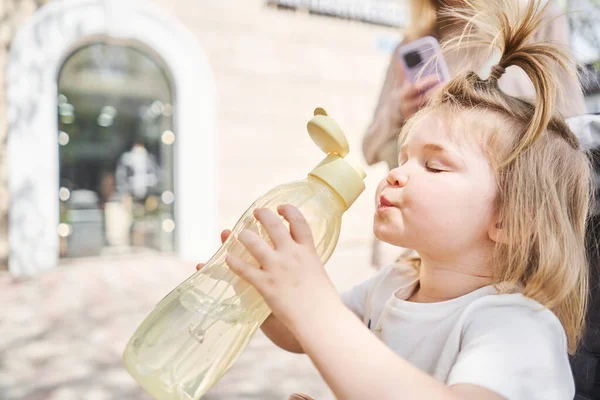 Büyüleyici küçük bir kız şehir parkındaki bir spor şişesinden su içiyor. Sıcak bir yaz gününde şişeden su içen bir çocuğun yakın plan portresi. Yüksek kalite fotoğraf