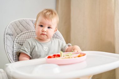 Tatlı çocuk sağlıklı makarna ve sebze buğulama yiyor. 10 aylık güzel bir kızın portreleri. Bebek bebekler için özel bir sandalyede oturuyor. Yüksek kalite fotoğraf