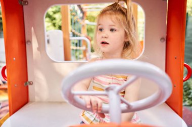 Oyun oynayan küçük bir kız, çocuk parkında direksiyon tutan bir araba şoförü. Güneşli bir yaz akşamı, eğlenceli bir çocukluk. Yüksek kalite fotoğraf