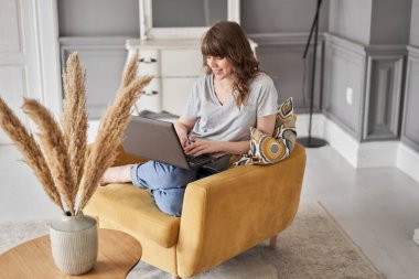 Evdeki oturma odasında kanepede otururken siyah dizüstü bilgisayar kullanan güzel, neşeli bir kız. Genç bir kadın iletişim ve iş için dizüstü bilgisayar kullanıyor. Evet.