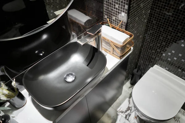 A black sink with a silver faucet next to an oval mirror and a shelf with hand paper towels. Close-up of an elegant faucet in the bathroom sink next to stylish decorations. High quality photo