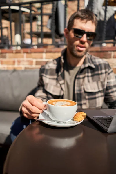 A man drinks coffee, holds a cup of hot Peanut latte in his hand. The table in the coworking space of the coffee shop. A cup of coffee on the veranda on a sunny day. Coffee break at the cafe. Close-up