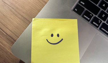 Laptoptaki sarı yapışkan notta gülen surat. Laptopta üzerinde gülen yüz olan yapışkan bir not. Takdir göstergesi ya da özür dileme.
