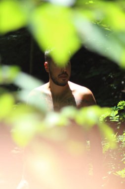 Genç, dövmeli ve kaslı bir adam Zürih 'te güneşli bir günde yemyeşil yapraklar tarafından çerçevelenmiş duruyor. Şehir manzarası sahneye canlı bir şehir dokunuşu katıyor.