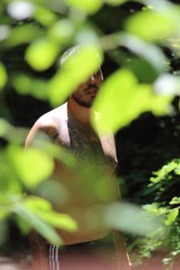 Genç, dövmeli ve kaslı bir adam Zürih 'te güneşli bir günde yemyeşil yapraklar tarafından çerçevelenmiş duruyor. Şehir manzarası sahneye canlı bir şehir dokunuşu katıyor.