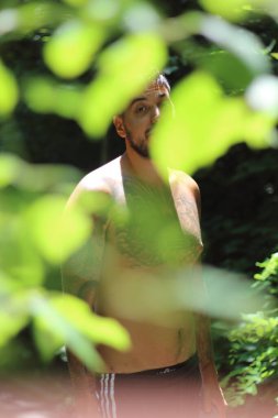 Genç, dövmeli ve kaslı bir adam Zürih 'te güneşli bir günde yemyeşil yapraklar tarafından çerçevelenmiş duruyor. Şehir manzarası sahneye canlı bir şehir dokunuşu katıyor.