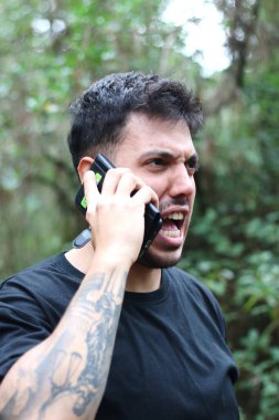 A frustrated young man yells into his cell phone during an intense phone call, standing amidst a peaceful forest clipart