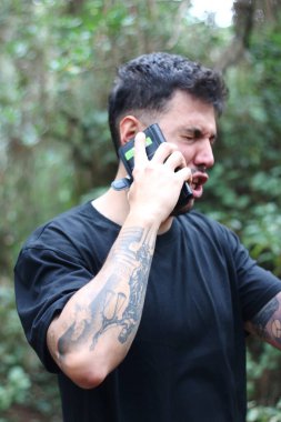 A frustrated young man yells into his cell phone during an intense phone call, standing amidst a peaceful forest clipart