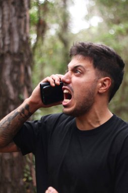 A frustrated young man yells into his cell phone during an intense phone call, standing amidst a peaceful forest clipart