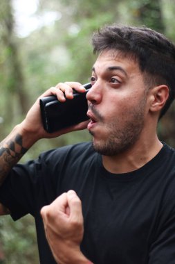 A frustrated young man yells into his cell phone during an intense phone call, standing amidst a peaceful forest clipart