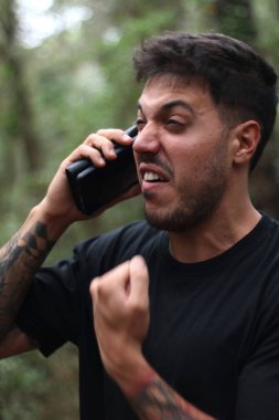 A frustrated young man yells into his cell phone during an intense phone call, standing amidst a peaceful forest clipart