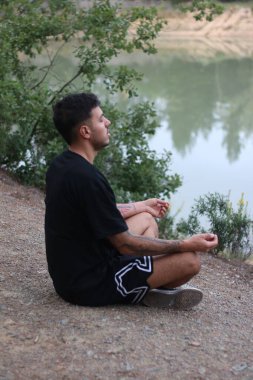 Genç bir adam sakin bir gölün kenarında huzur içinde oturur, sakin ve düşünceli bir şekilde meditasyon yapar. Huzurlu sahne, vücut ve doğa arasındaki uyumu yakalar.