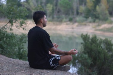 Genç bir adam sakin bir gölün kenarında huzur içinde oturur, sakin ve düşünceli bir şekilde meditasyon yapar. Huzurlu sahne, vücut ve doğa arasındaki uyumu yakalar.